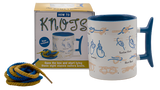 A white mug with a blue inside and a blue handle that has illustrations on how to tie knots on it. The side of the mug facing the camera has instructions on tying a bowline, a tautline hitch, and a sheet bend. There is a box for the mug that shows an image of the mug on it along with text that states, "How to Knots. Rope included! Open the box and start tying these eight classic sailors knots." There is some rope next to the mug to practice tying knots with, too.