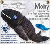 A string doll of a whale. We can see two versions of it, one with a black body with a blue eye, and another with a blue body and a black eye. Both have white water coming out of their blow holes. Text on the image reads, "Moby reminds you to surface every once in a while and take a deep breath."