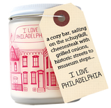 A candle with an illustration of different houses with a bird flying over them, stating "I love Philadelphia." The illustration has a sketchy style done in pink. There is a tag on the candle that reads, "A cozy bar, sailing on the Schuylkill, cheesesteak with grilled onions, historic streets to museum steps...I love Philadelphia."