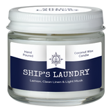 A white candle in a container. It has a white and blue label that says, "Hand poured coconut wax candle. Ship's Laundry. Lemon, clean linen & light musk."