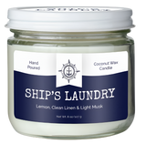 A white candle in a container. It has a white and blue label that says, "Hand poured coconut wax candle. Ship's Laundry. Lemon, clean linen & light musk."