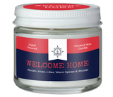 A candle in a glass container. There is a red and blue label on it that reads, "Hand poured coconut wax candle. Welcome home. Roses, irises, lilies, spices & woods."