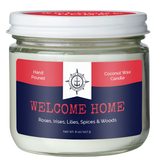 A candle in a glass container. There is a red and blue label on it that reads, "Hand poured coconut wax candle. Welcome home. Roses, irises, lilies, spices & woods."