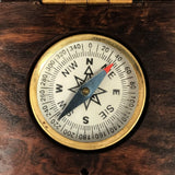 A close-up of a vintage brass compass with a white dial, black degree markings, and a blue needle pointing north, set inside a dark brown wooden box.
