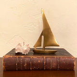 A small brass sailboat figurine sits on top of an old brown leather-bound book, with a white seashell beside it against a textured cream wall.