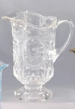 A clear glass pitcher with ornate embossed floral and scrollwork patterns, featuring a wide flared base and a curved handle, shown against a neutral gray background.