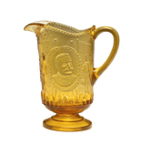 An amber yellow glass pitcher with embossed portrait and decorative motifs on the front, set against a neutral gray background.