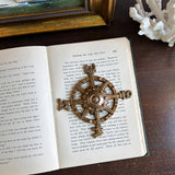 Brass ship's wheel compass paperweight on an open book with a wooden surface and decorative items in the background.