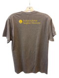 A dark gray heather t-shirt hanging on a clear plastic hanger against a textured gray wall. The back of the shirt features a yellow printed logo and text that reads 'Independence Seaport Museum'.