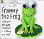 A frog string doll. It has a green body and black and white eyes with a black smile. It is sitting on a green lily pad with a daisy on it. There is text on the lefthand side that reads, "Franny the frog helps you leap over whatever is holding you back."