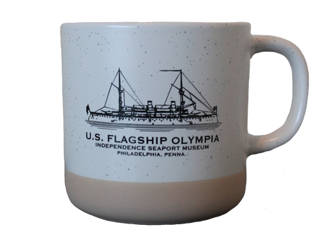 A white mug with black speckles and a tan bottom with a depiction of the Olympia on it. There is black text underneath the Olympia which reads, "U.S. Flagship Olympia. Independence Seaport Museum. Philadelphia, Penna." 