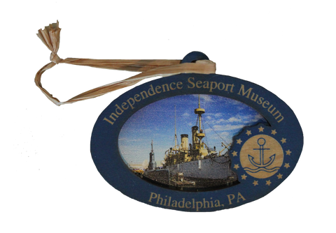 A wooden ornament that shows an image of the Cruiser Olympia on it, along with text that says, "Independence Seaport Museum. Philadelphia, PA," along with the ISM logo, which is an anchor floating on some waves encompassed in a circle and surrounded by stars. 