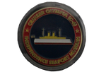 A challenge coin that has a simplified illustration of the U.S. Olympia on it. The boat has a gold top, a white base, and a red bottom, with a small U.S. flag on the back. It is blue around the ship and then red around the rest of the coin. Around the coin, it reads, "Cruiser Olympia (C-6). Independence Seaport Museum."