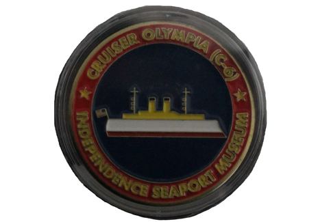 A challenge coin that has a simplified illustration of the U.S. Olympia on it. The boat has a gold top, a white base, and a red bottom, with a small U.S. flag on the back. It is blue around the ship and then red around the rest of the coin. Around the coin, it reads, "Cruiser Olympia (C-6). Independence Seaport Museum."