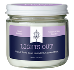 A candle with a blue and purple label on the front of it with text that reads, "Hand poured coconut wax candle. Lights out. Wood, tonka bean, lavender, & coconut milk." There is a logo in the center of the candle, depicting an anchor surrounded by a compass-like shape, with the words, "Fair wind candle company" around the base of it. 