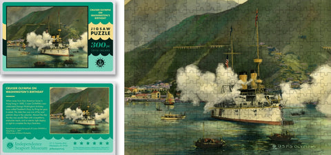 A puzzle of Olympia celebrating Washington's birthday. The Olympia is shown in the water, with golden smokestacks, a white base, and a red bottom, firing its cannons. There is smoke from it firing the cannons. Next to the ship is a mountain with different homes on it. There are a few others, smaller boats in the water next to the Olympia. The puzzle box is shown, it is teal blue with a picture of the completed puzzle on it. 