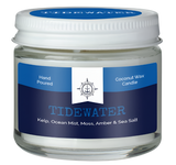 A coconut wax candle in a jar. There is a blue label on it that reads, "Hand poured coconut wax candle. Tidewater. Kelp, ocean mist, moss, amber & sea salt."  