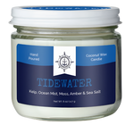 A coconut wax candle in a jar. There is a blue label on it that reads, "Hand poured coconut wax candle. Tidewater. Kelp, ocean mist, moss, amber & sea salt."  