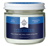 A coconut wax candle in a jar. There is a blue label on it that reads, "Hand poured coconut wax candle. Tidewater. Kelp, ocean mist, moss, amber & sea salt."  